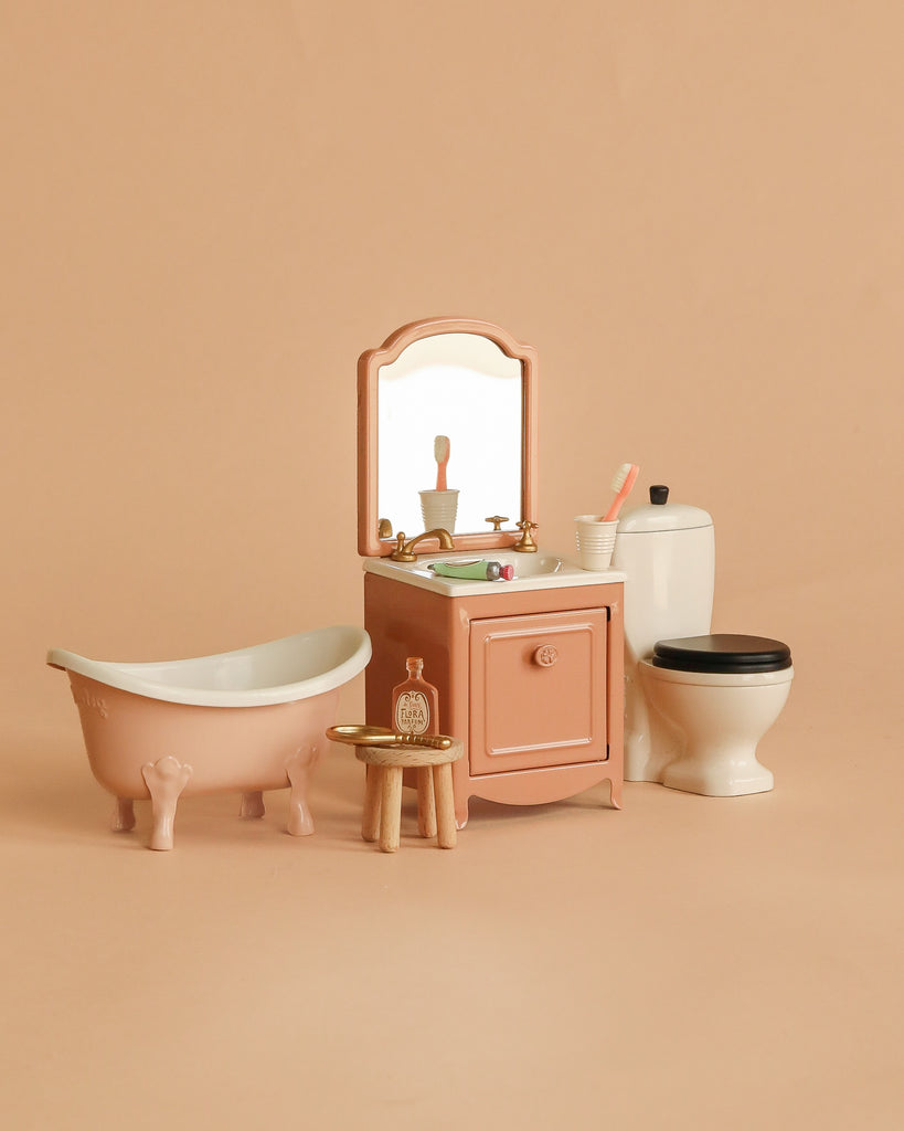 The Maileg Bathroom Set includes a bathtub, vanity with mirror, sink, toothbrush, soap, stool with bottles, and toilet in soft pink and white—ideal for displaying Maileg mouse furniture against a beige background.