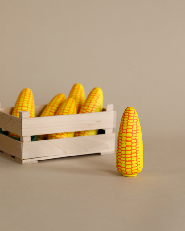 A wooden crate filled with several Erzi Corn on the Cob Pretend Foods with a prominent yellow and white patterned cob standing upright in front of it on a plain background.