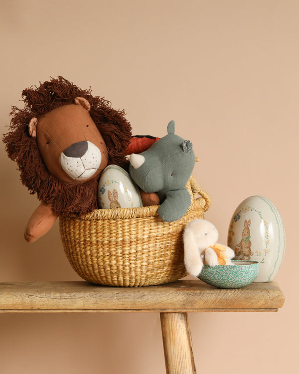 A handmade Easter Basket holds plush toys such as a Maileg Rhino Safari and lion, with an Easter Egg Set displaying animal illustrations. Beside it, a ceramic bowl features a bunny toy and matching eggs on the wooden bench. Beige background.