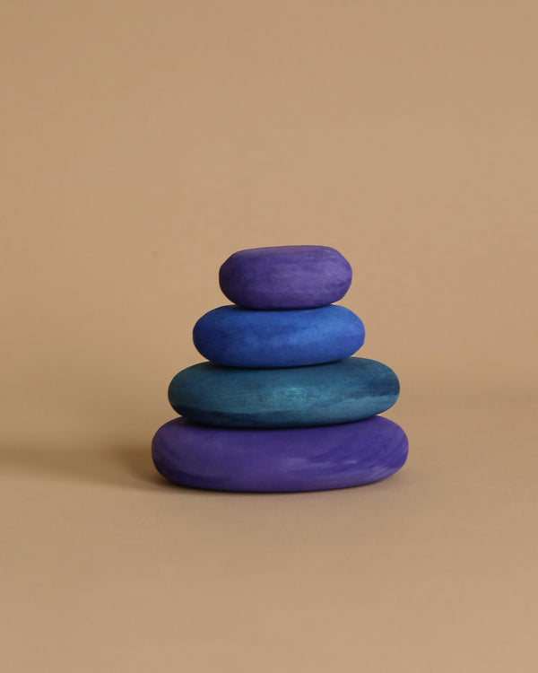 The Grimm's Dream Pebbles Stacker features a collection of four smooth, organic round stone shapes in varying shades of blue and purple against a beige background. Each piece is colored with non-toxic water-based stains and arranged from largest at the bottom to smallest at the top, creating a balanced and symmetrical tower.