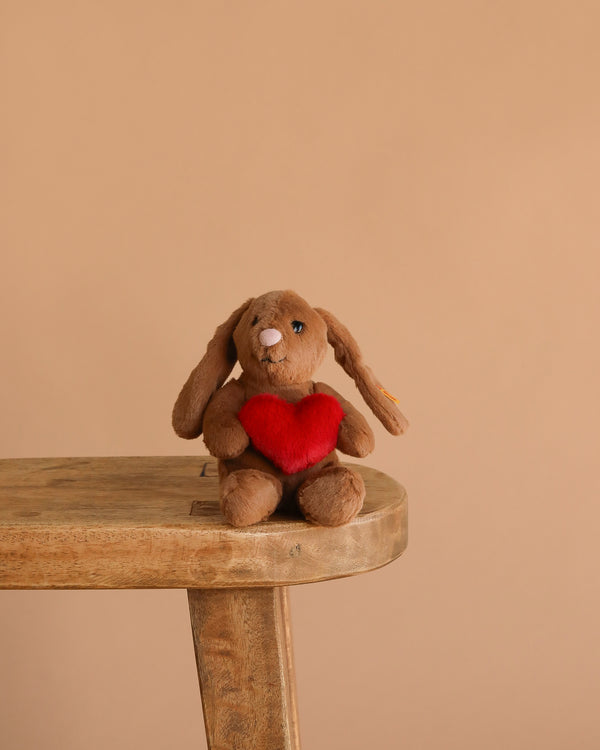 Steiff Hoppie Rabbit – Love, a plush stuffed animal holding a red heart, sits on a wooden stool against a plain beige background—a perfect gift for Steiff fans.