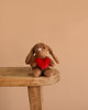 Steiff Hoppie Rabbit – Love, a plush stuffed animal holding a red heart, sits on a wooden stool against a plain beige background—a perfect gift for Steiff fans.