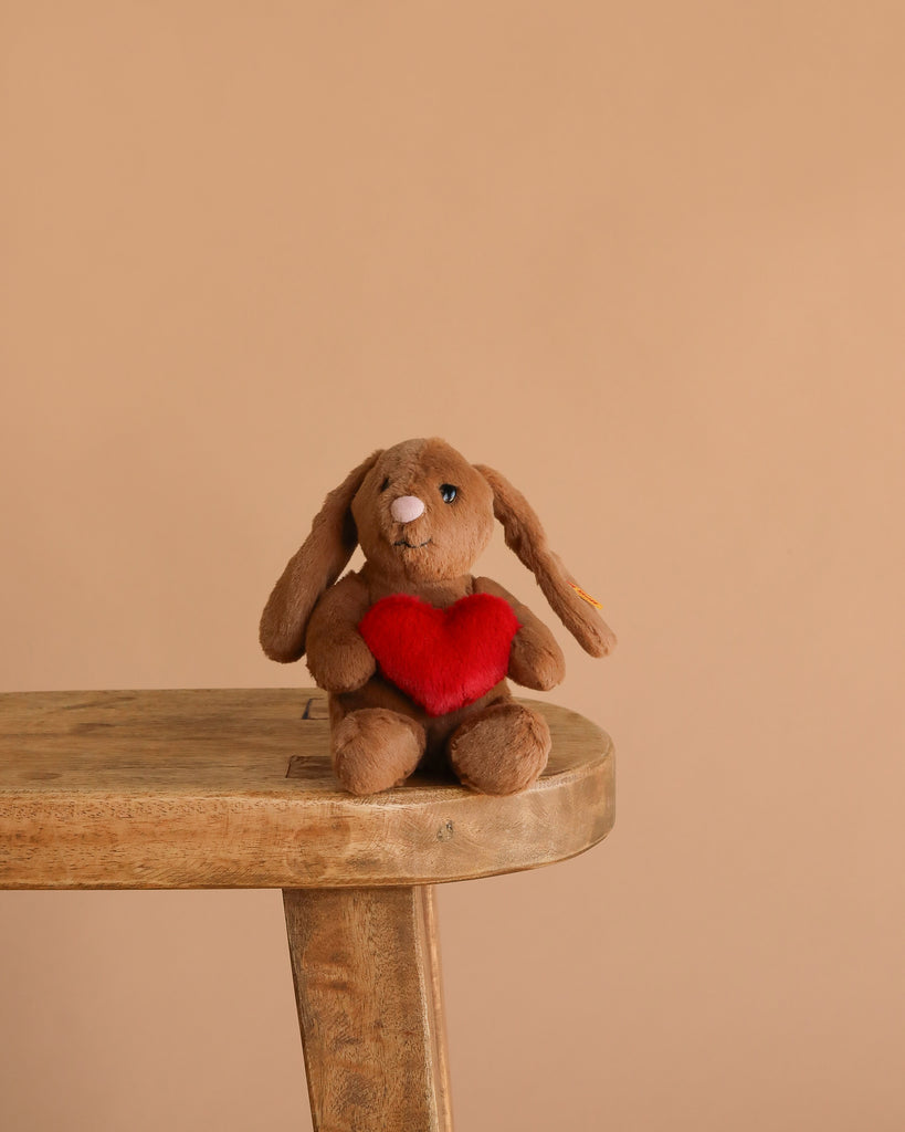 Steiff Hoppie Rabbit – Love, a plush stuffed animal holding a red heart, sits on a wooden stool against a plain beige background—a perfect gift for Steiff fans.