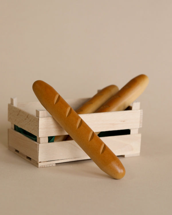 Two Erzi Baguette Pretend Foods with golden crusts, handcrafted in Germany, placed in a small wooden crate against a neutral background.