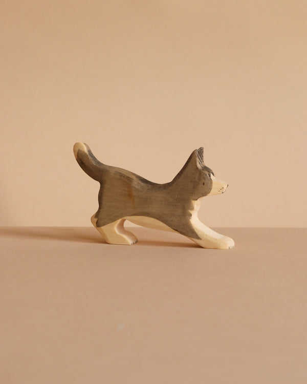 The Handmade Wooden Border Collie is a small, handcrafted dog figurine with a raised tail, painted in brown and beige plant-based stains. It stands on a light brown surface with a matching background.