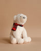 The Mon Ami - North Polar Bear Plush Toy, wearing a red and black plaid scarf, sits on a beige surface against a plain beige background.
