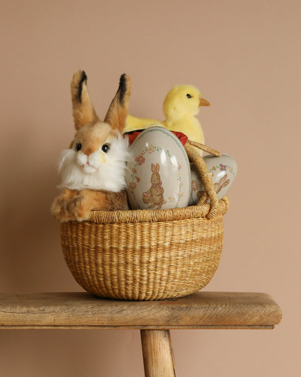 An Easter Basket, handmade in Bolgatanga, Ghana, sits on a wooden stool. Inside are a plush rabbit, Maileg Easter Egg, and a yellow chick plush amidst decorative eggs with bunny designs against a soft beige background.