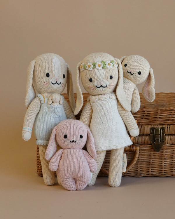 Four adorable Cuddle + Kind Bunny hand-knit rabbit dolls of various sizes posed in front of a wicker basket, each with a unique, friendly smile and outfit, on a neutral background.