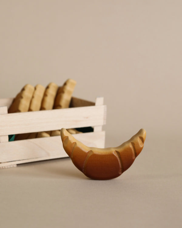 A realistic Erzi Croissant Pretend Food model in the foreground with a wooden crate filled with similarly styled baguettes in the background, all set against a beige backdrop.