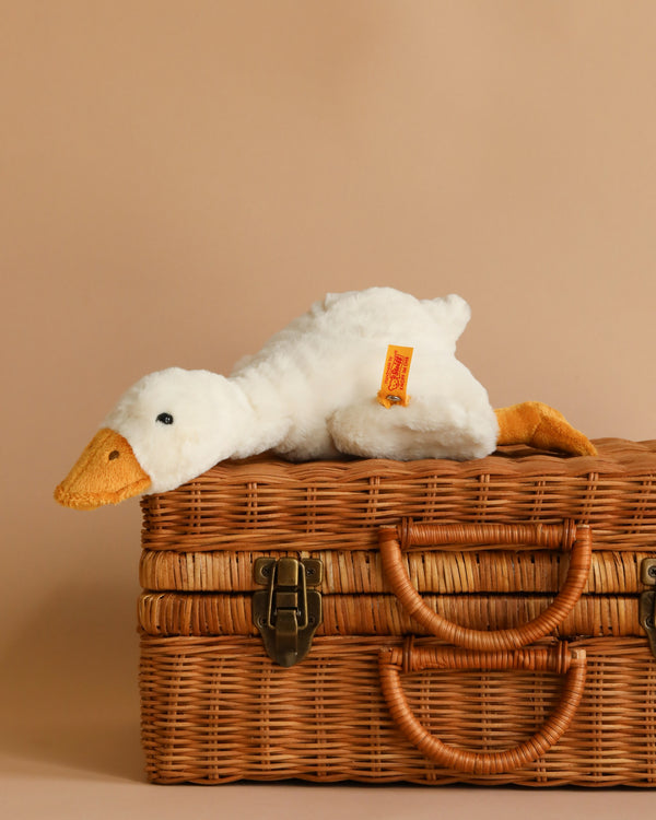 A white plush goose stuffed animal lying on top of a wicker basket with a latch, against a soft beige background. The toy features a visible Steiff Button in Ear tag on its side.