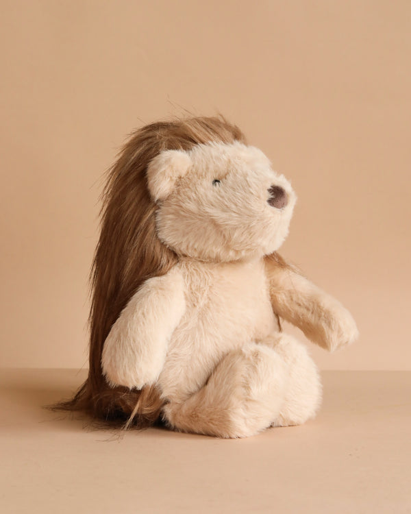 The Mon Ami - Harlow Hedgehog Plush Toy, featuring soft, light brown fur and long, dark brown hair, sits upright against a beige background and faces slightly to the right.