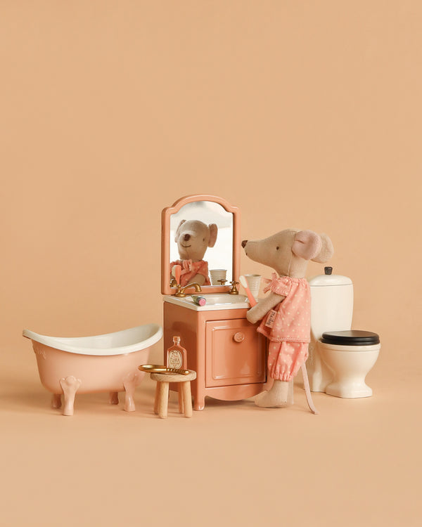 The Maileg Big Sister Bathroom Set features a mouse in pink pajamas at a peach vanity with mirror, matching bathtub, and white toilet—charming accessories for imaginative play against a beige backdrop.