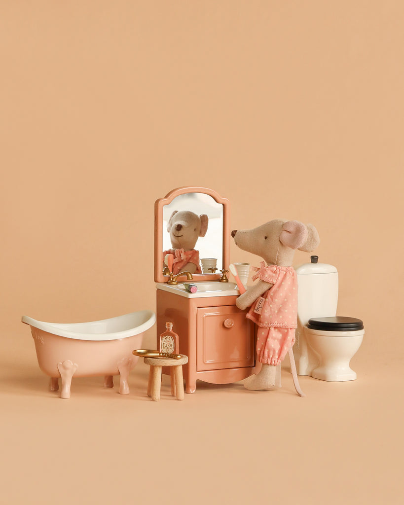 The Maileg Big Sister Bathroom Set features a mouse in pink pajamas at a peach vanity with mirror, matching bathtub, and white toilet—charming accessories for imaginative play against a beige backdrop.