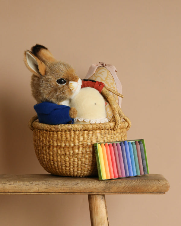 An Easter Basket on a wooden stool holds a Blue Jacket Bunny stuffed animal, a soft white ball, and a patterned gift. In front sits a box of multicolored crayons, all against a plain beige wall.