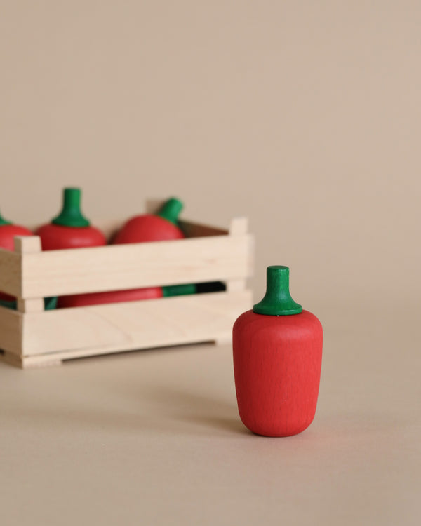 A handcrafted wooden toy Erzi Red Pepper Pretend Food with a green stem stands in the foreground, with more toy Erzi Red Pepper Pretend Food in a miniature wooden crate blurred in the background. The neutral-toned background highlights the vibrant red and green.