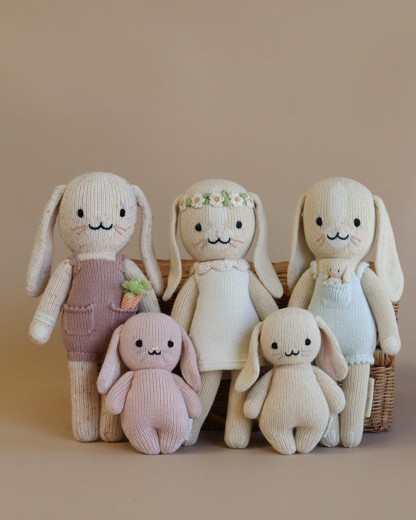 Five Cuddle + Kind Bunny hand knit stuffed animals in pastel colors posing together: two rabbits, two smaller creatures, and one in a floral crown, all near a basket.