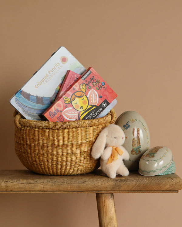 A handmade Easter Basket on a wooden stool contains a Stockmar Triangular Coloured Pencil Set and two boxes of organic tea. A small plush bunny toy is beside it, while two egg-shaped containers featuring rabbit illustrations are nearby against a plain brown background.