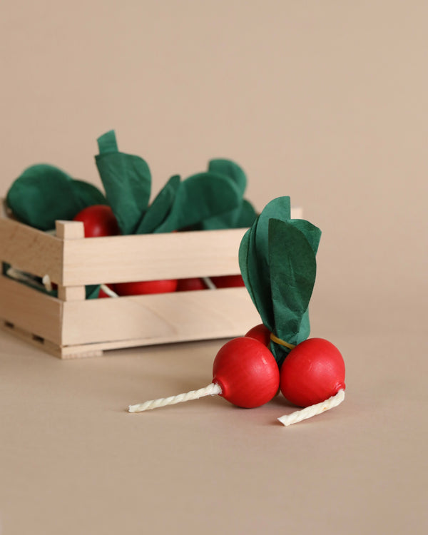 Two realistic Radish Bunch Pretend Foods handcrafted from red and green paper displayed in front of a small wooden crate on a beige background.