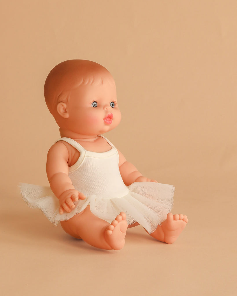 The Minikane Vintage Baby Doll (13") - Lily with Clothing, made in Spain, wears a white tutu dress and sits on a beige background, featuring blue eyes, rosy cheeks, and short molded brown hair facing slightly to the right.