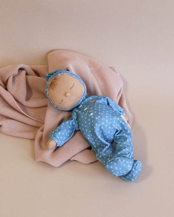 A Olli Ella Dozy Dinkums - Bug from the Limited-edition Daydream Collection, wearing a blue outfit with white stars, lying on a beige blanket, set against a plain light beige background. The doll has a serene expression.