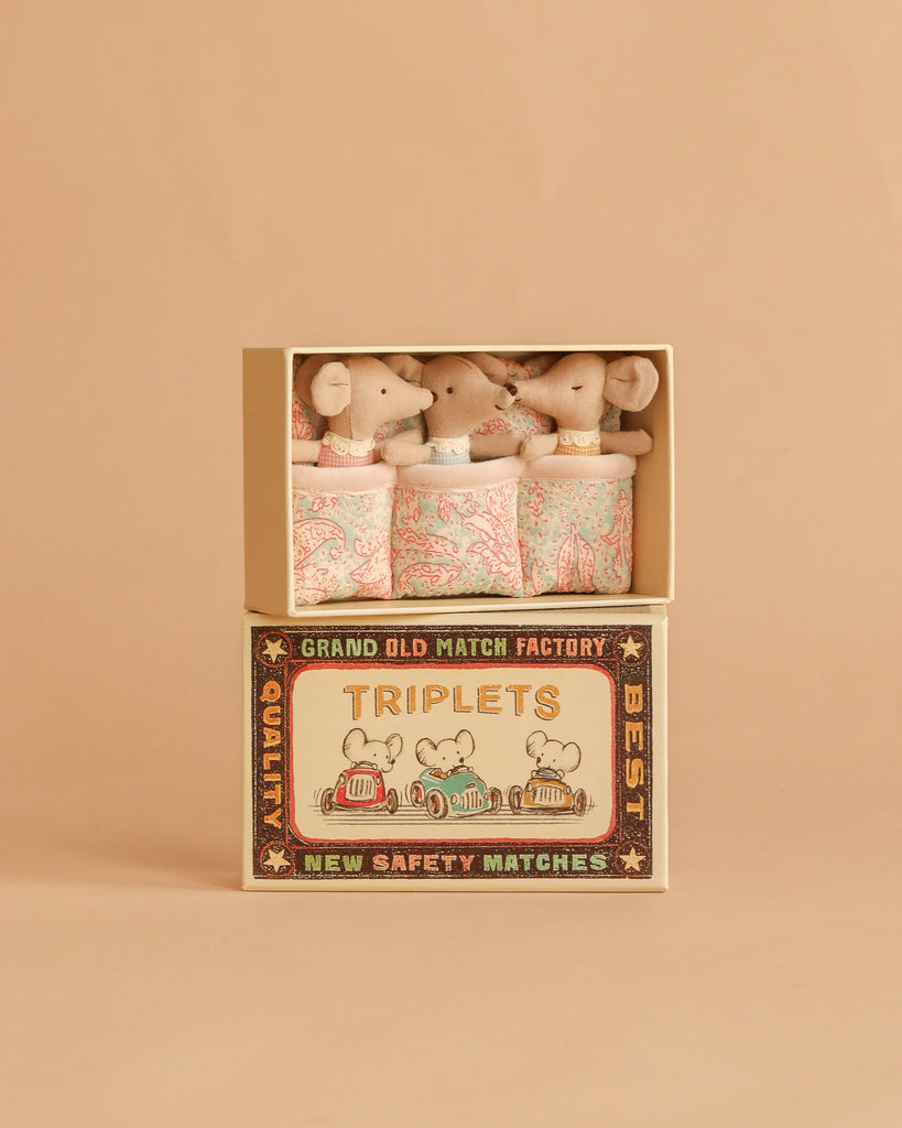 Three baby mice snuggle in patterned blankets inside the Maileg Triplets, Baby Mice in Matchbox - Checker set, featuring a soft beige background and a matchbox decorated with three cartoon mice in colorful pajamas.