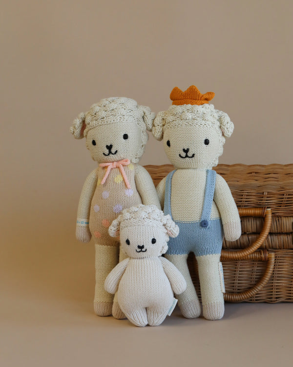 Two Cuddle + Kind Lamb Stuffed Animals, one in pink and blue attire with a baby version, and the other in blue overalls and an orange hat, sit beside a woven basket on a beige background.