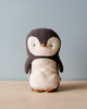 The Maileg Penguin Stuffed Animal - Small, crafted from soft fabrics, is displayed on a wooden surface against a gentle blue background. This endearing design, featuring a predominantly dark grey color with a cream-colored belly and face along with tiny orange feet and beak, is sure to appeal to people of all ages.