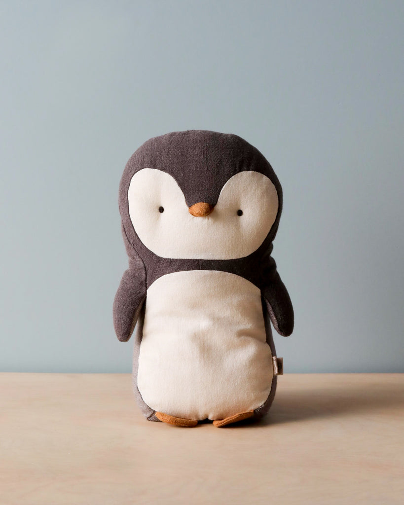 The Maileg Penguin Stuffed Animal - Small, crafted from soft fabrics, is displayed on a wooden surface against a gentle blue background. This endearing design, featuring a predominantly dark grey color with a cream-colored belly and face along with tiny orange feet and beak, is sure to appeal to people of all ages.