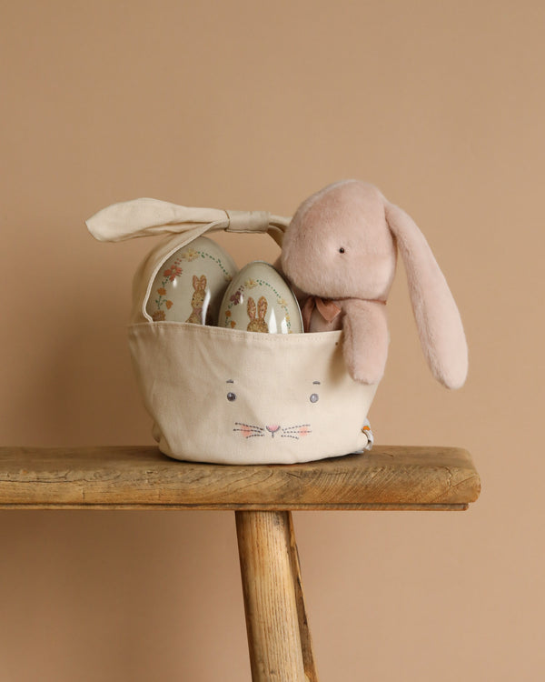 Against a beige backdrop, a Maileg Plush Bunny leans on an Easter Basket with a bunny face design, perched on a wooden stool. Inside the basket sits an Easter Egg Set adorned with bunny illustrations.