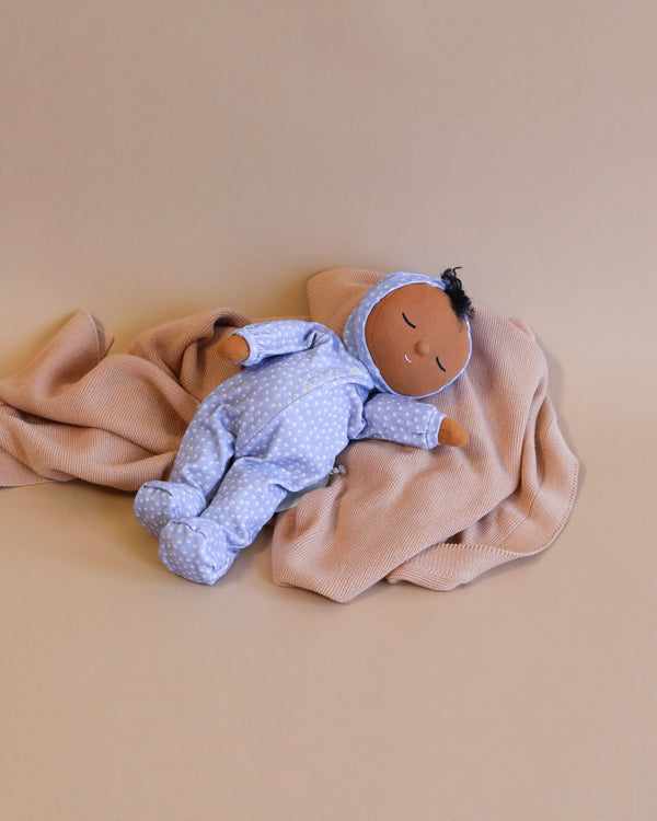 A soft fabric doll from the limited-edition Daydream Collection with dark skin and black hair, dressed in a blue and white polka dot onesie, resting on a beige blanket against a light brown Olli Ella Dozy Dinkums - Squeak.