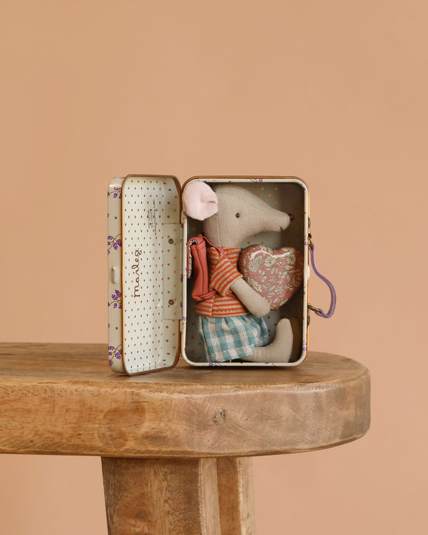 The Maileg Big Sister On the Go Suitcase Set features a small mouse in a striped shirt and plaid shorts, sitting inside an open suitcase on a wooden stool, hugging a heart pillow against a plain beige background.