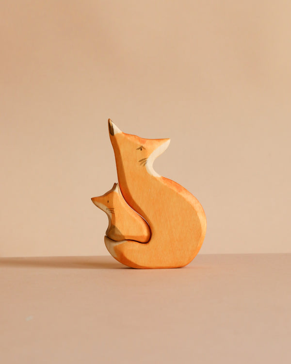 The Handmade Wooden Snuggling Foxes set features two minimalistic fox figurines, one large and one small, hand crafted from sustainable hardwood and finished with natural, non-toxic stains against a beige background.