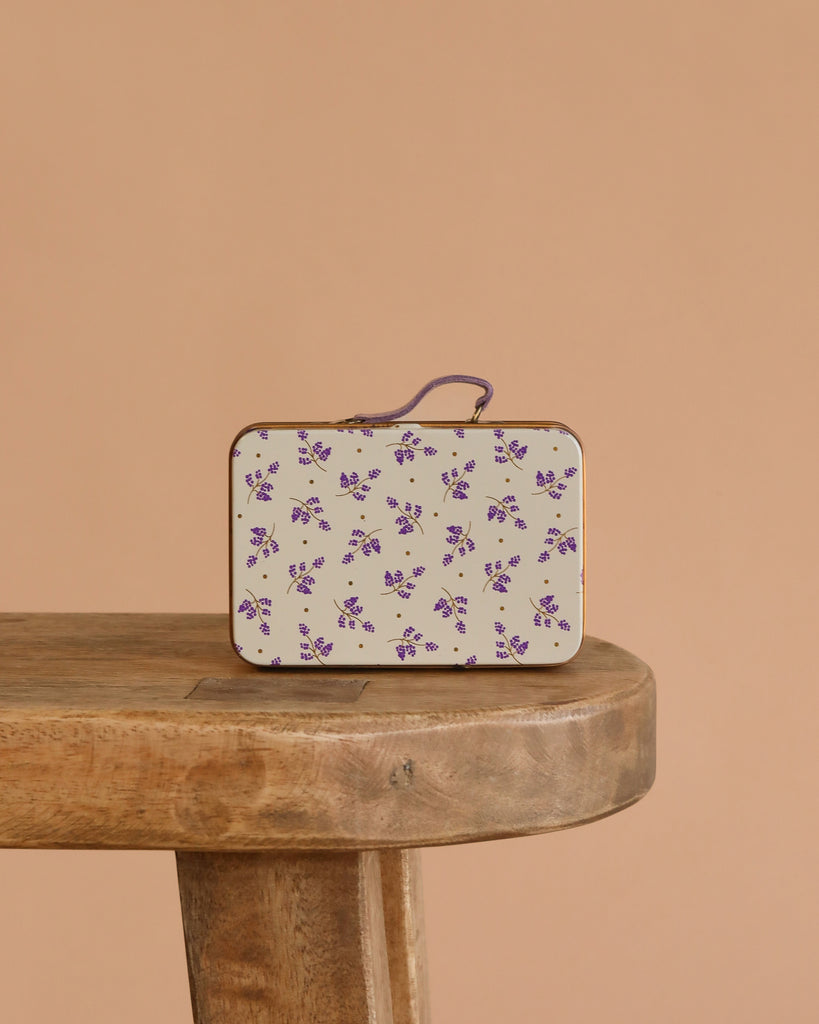 The Maileg Big Sister On the Go Suitcase Set, featuring a small rectangular case with a white and purple floral design, rests on a wooden bench against a plain beige background.
