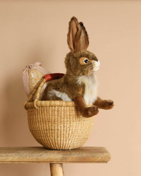 A Blacktail Rabbit stuffed animal rests in a Handmade Easter Basket of elephant grass, on a wooden stool against a beige background. A pink patterned fabric pouch is partially visible behind the rabbit.