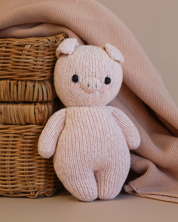 A handmade knitted Cuddle + Kind Baby Piglet plush toy, crafted from Peruvian cotton yarn, standing in front of a wicker basket, partially covered by a soft beige blanket. The pig has a smiling face and
