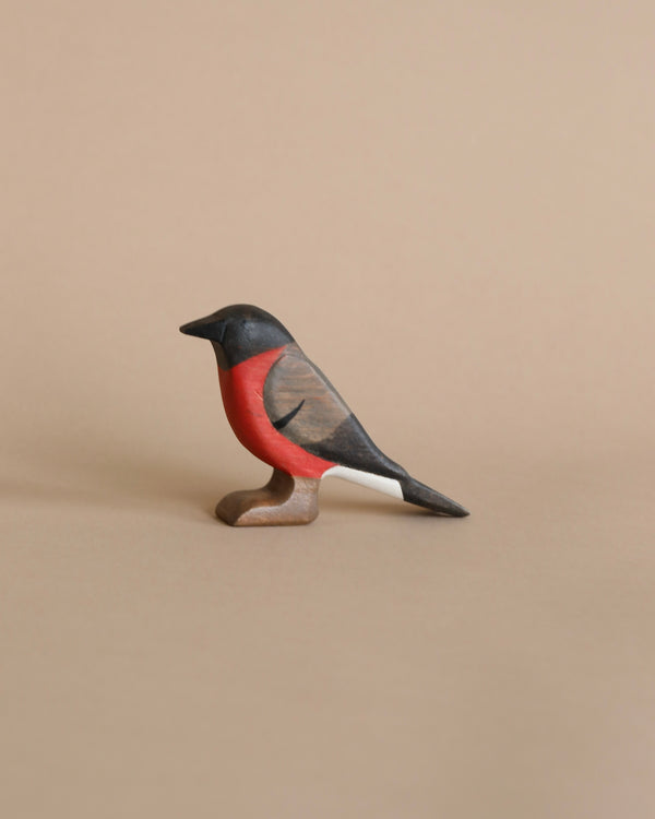 A Handmade Wooden Bullfinch Bird figurine painted with non-toxic water-based paint, featuring black, red, and white coloring on a light brown background. The bird is positioned sideways with its