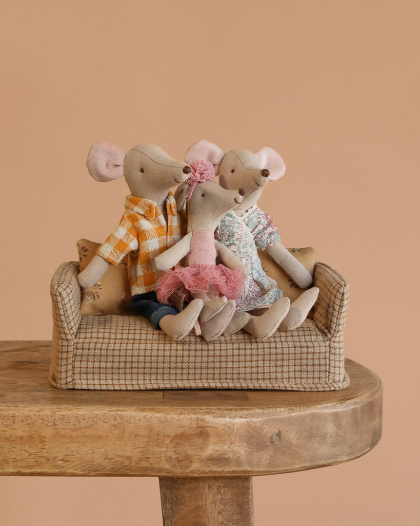 Three fabric ballerina mouse dolls in colorful clothes sit together on a patterned couch, on a wooden table with a beige background, highlighting the Maileg Family Room Set's whimsical charm.