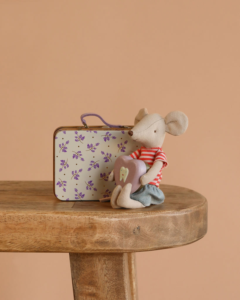 The Maileg Big Sister On the Go Suitcase Set features a small mouse in a red striped shirt and blue shorts holding a heart pillow, sitting beside a floral metal suitcase on a wooden table with a light brown background.