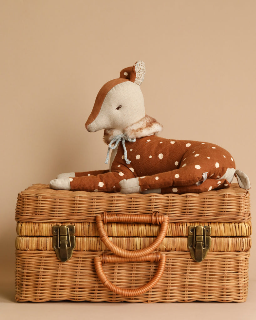 A Maileg Cosy Bambi Deer - Mint, featuring a brown body with white spots and adorned with a blue ribbon around its neck, rests on a neatly closed woven wicker suitcase with a leather handle. This charming piece is set against a neutral beige background, making it ideal for decoration or play.