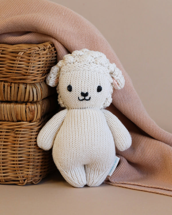 A Cuddle + Kind Baby Lamb, hand-knit from Peruvian cotton yarn, with a smiling face sits beside a wicker basket, partially covered by a soft pink blanket against a beige background.