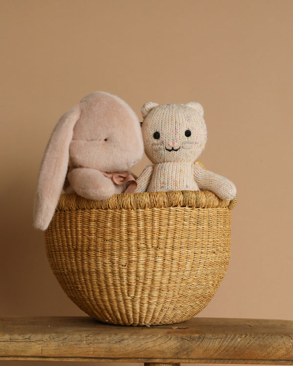 A handmade Easter Basket on a wooden surface holds a plush bunny with long ears and a knitted smiling bear toy, crafted from Peruvian cotton yarn. The plain beige background adds to the warm and cozy atmosphere.