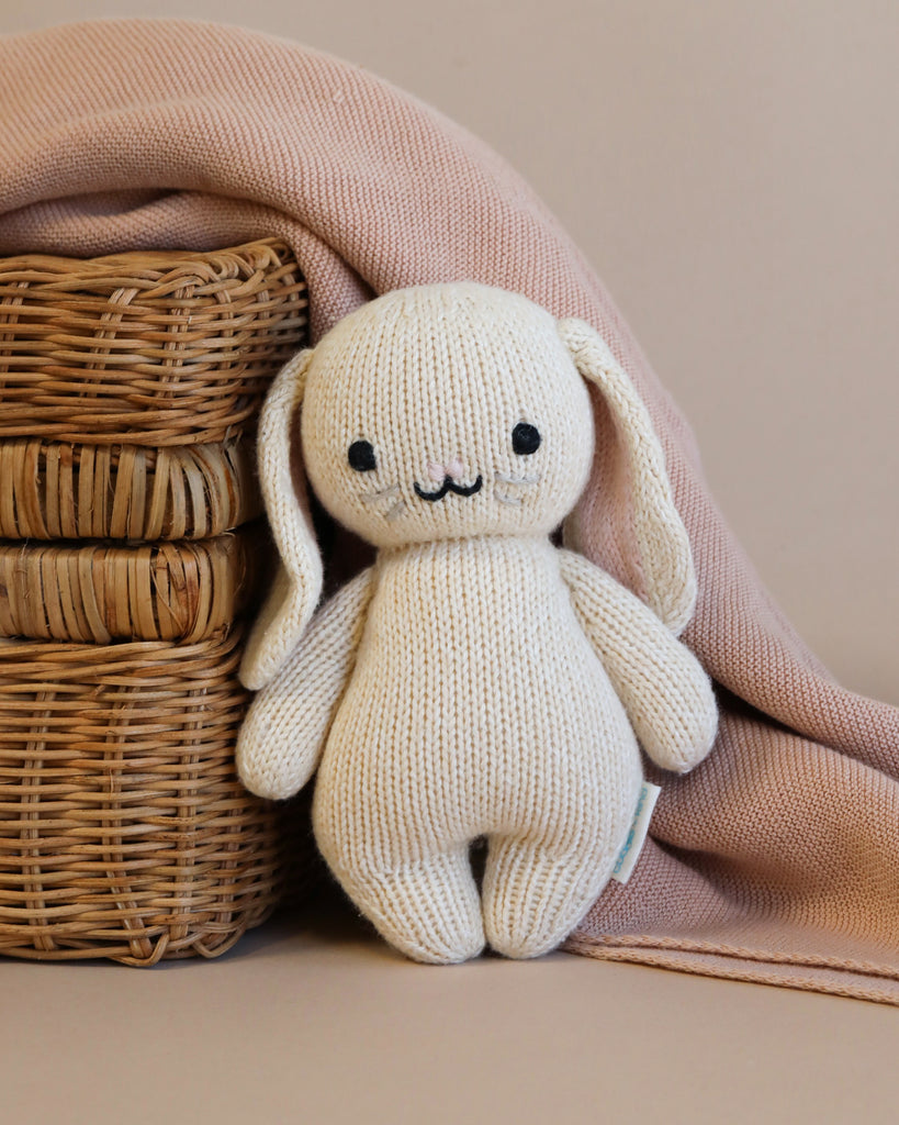 A Cuddle + Kind Baby Bunny toy with long floppy ears sitting next to a wicker basket, partially covered by a soft pink cloth on a beige background.