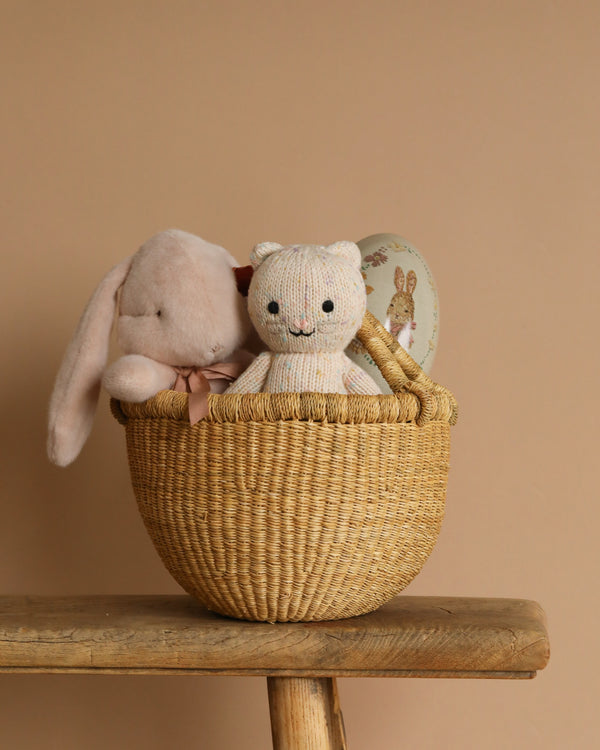 An Easter Basket rests on a bench against a beige background, containing a Maileg plush bunny, a knitted bear toy, and a round box with an endearing bunny illustration.