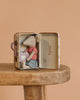 The Maileg Big Sister On the Go Suitcase Set features a small mouse in a red striped shirt and blue shorts, holding a tooth-shaped pillow while sitting inside an open suitcase atop a wooden stool, set against a plain peach background.