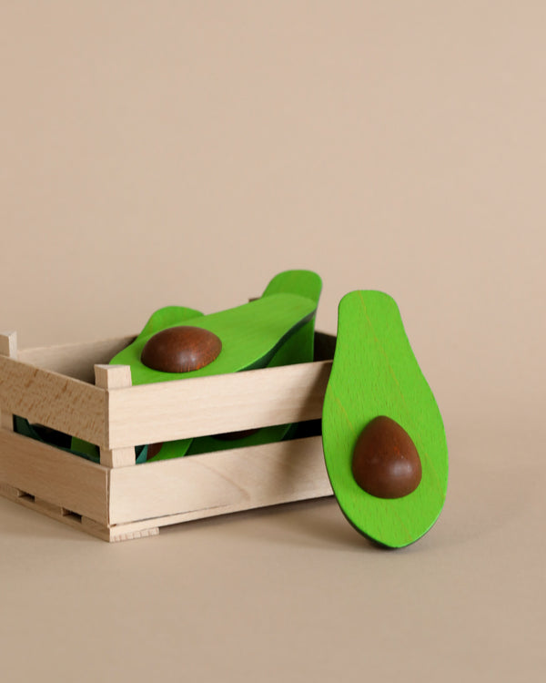 A handcrafted Erzi Avocado Half Fruit Pretend Food with a removable brown pit nestled in a small, slatted wooden crate against a neutral beige background.