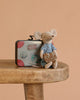 A small Maileg LouiMax mouse in a striped shirt and blue pants sits on a wooden table, holding a patterned pillow, with the Maileg LouiMax On the Go Suitcase Set featuring travel stickers placed behind. The background is light brown.