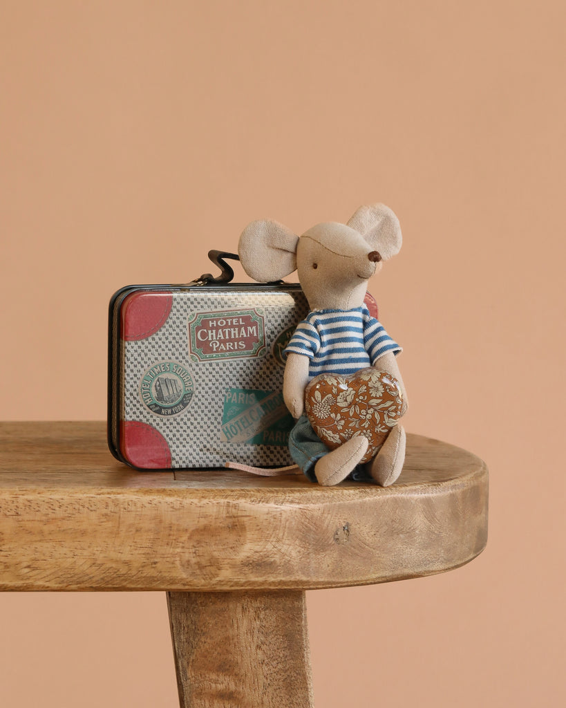 A small Maileg LouiMax mouse in a striped shirt and blue pants sits on a wooden table, holding a patterned pillow, with the Maileg LouiMax On the Go Suitcase Set featuring travel stickers placed behind. The background is light brown.