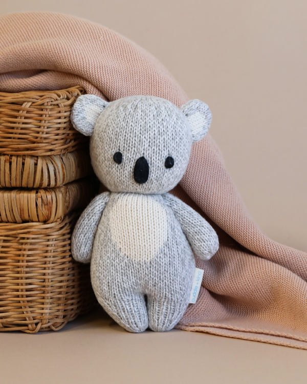 A hand-knit gray and white teddy bear, crafted from Peruvian cotton yarn, sits beside a Cuddle + Kind Baby Koala, draped with a soft pink cloth against a beige background.