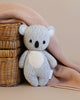 A hand-knit gray and white teddy bear, crafted from Peruvian cotton yarn, sits beside a Cuddle + Kind Baby Koala, draped with a soft pink cloth against a beige background.