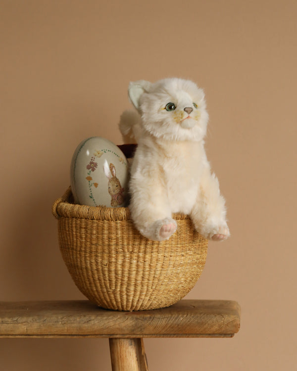 An Easter Basket, handmade in Bolgatanga, Ghana, holds a plush white cat stuffed animal and a decorative egg featuring a rabbit illustration. It sits on a wooden stool against a plain beige background.